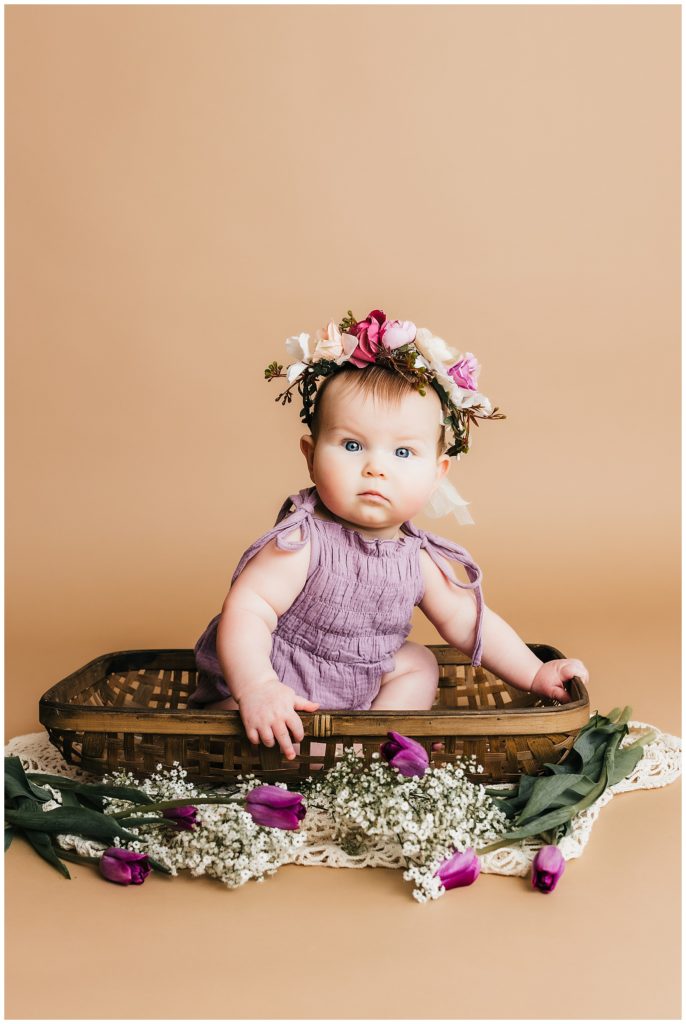 Baby girl sits in a woven basket wearing a floral crown among flowers for her sitter session