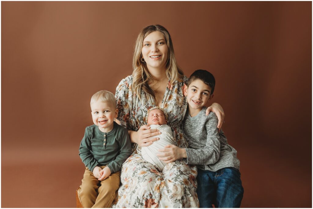 Mom with her two sons holding her swaddled baby girl on a chestnut backdrop during their Harrisburg newborn photography session