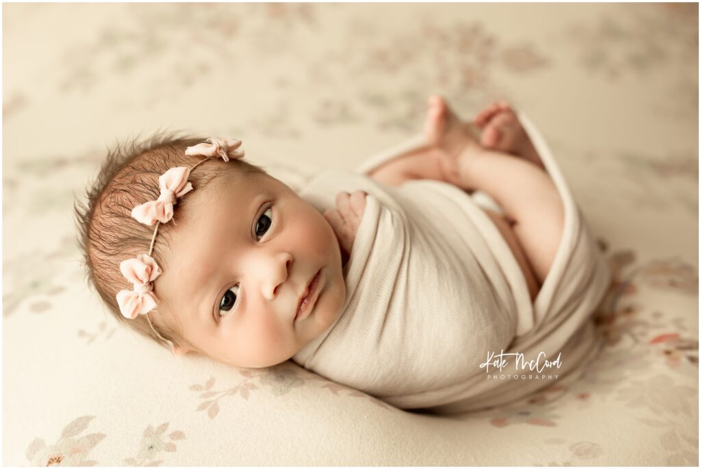 awake newborn baby girl with pink bow headband on a floral backdrop
