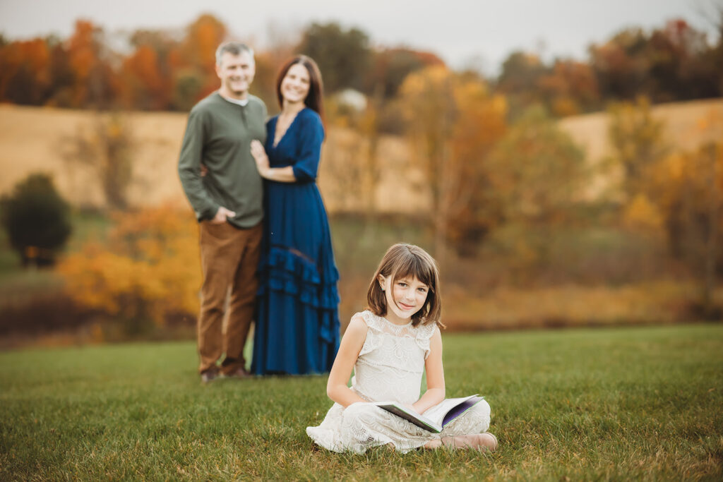 Parents smiling at child while she sits and reads her favorite Central PA library book outside  