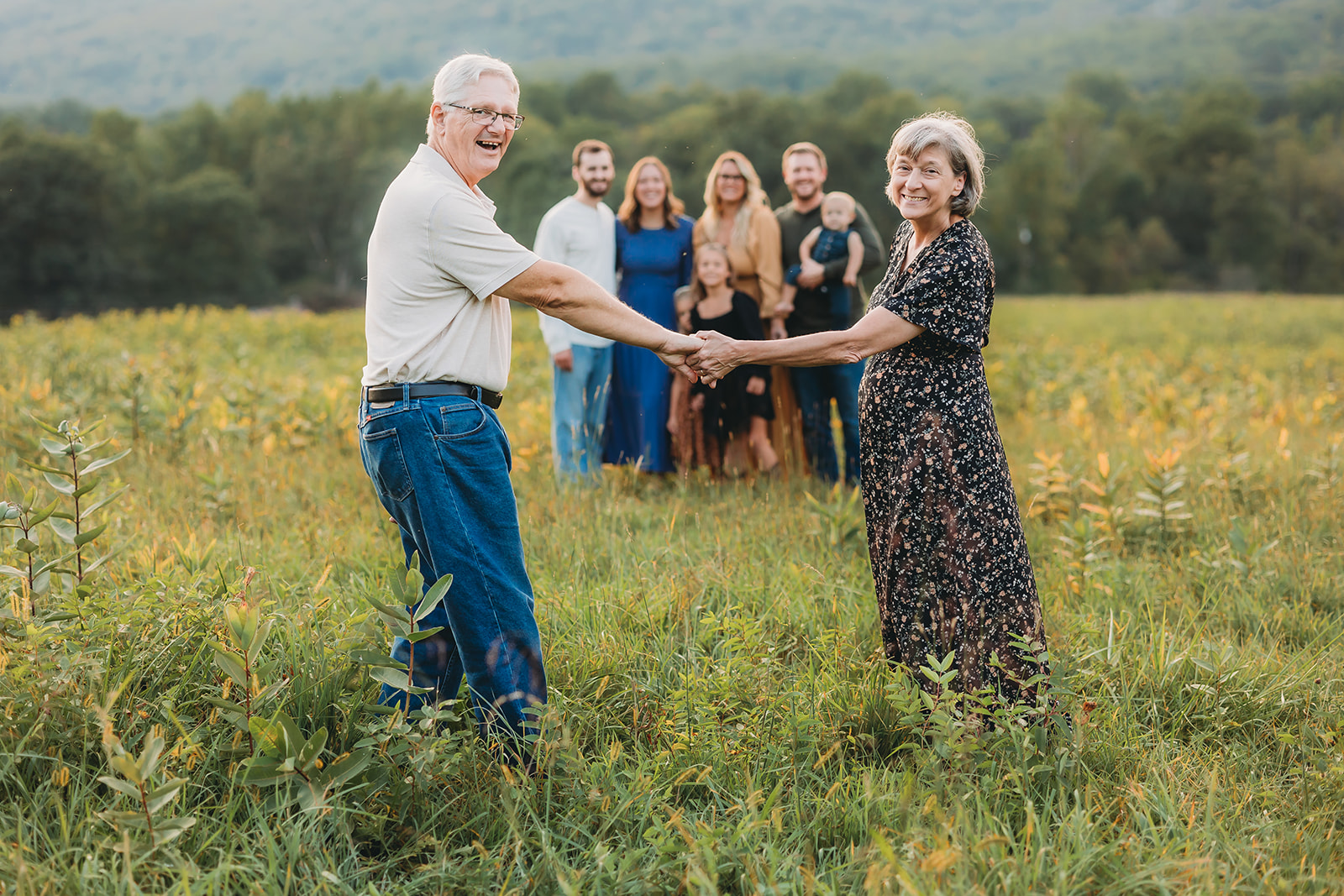 Husband and wife holding hands looking back at camera while their kids and grandkids smile in the background