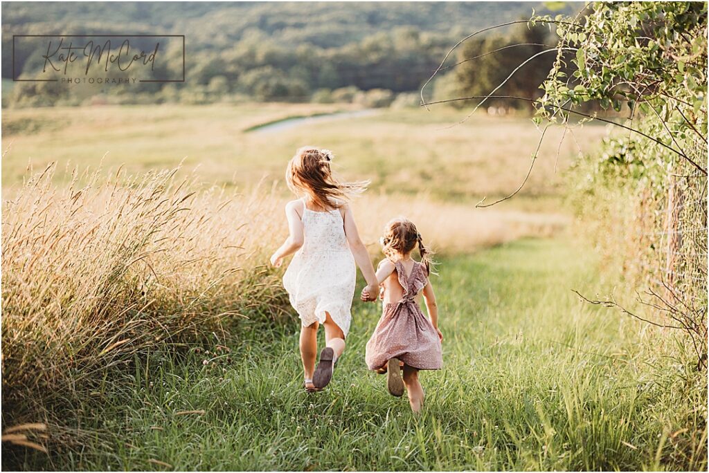 Sisters wearing dresses running hand in hand through a field at golden hour during a lifestyle candid photography session