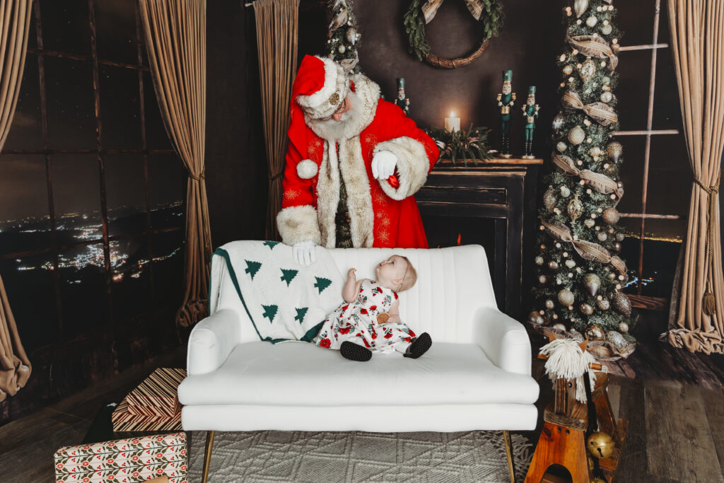 little girl in a christmas dress sitting on a couch looks up at santa as he sprinkles magic on her