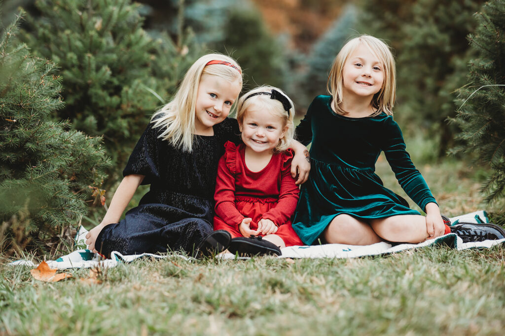 three sisters in holiday outfits smile big sitting on the floor at an etters christmas tree farm