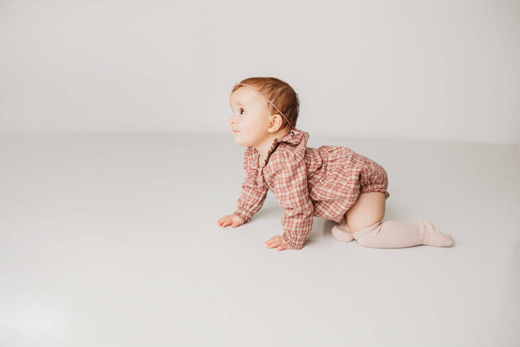 Baby in a plaid romper and knee highs crawls across a white backdrop and floor for milestone birthday photos