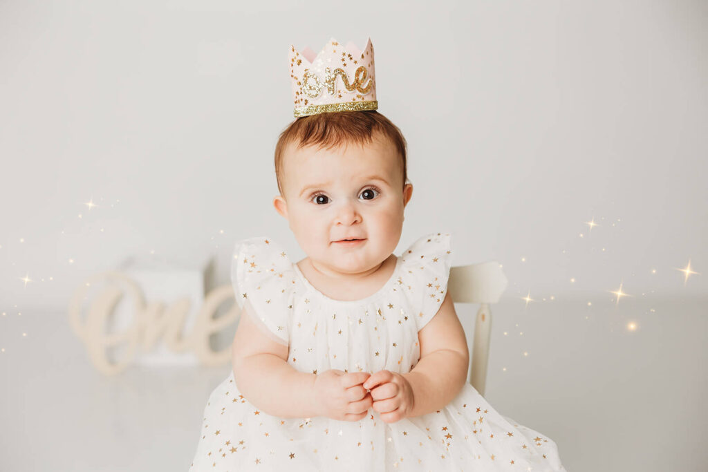 stella smiles with a sparkle in her eyes wearing a tulle star dress and a one crown on a simple white set for her first birthday portraits in mechanicsburg 