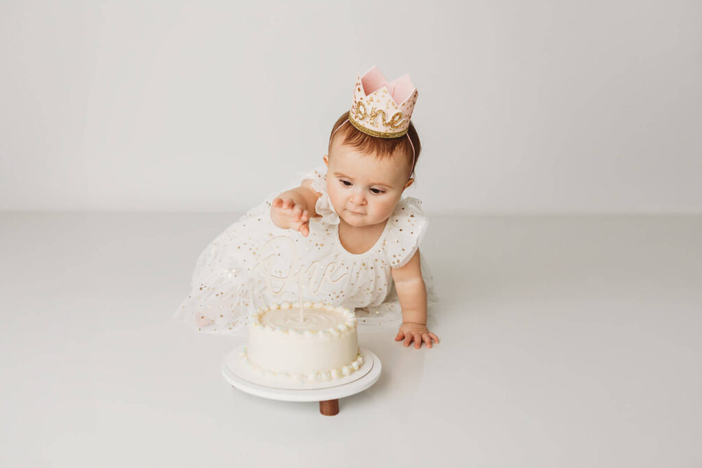One year old birthday girl in a tulle white dress with gold stars and a one crown reaches for her birthday cake in her cake smash session 