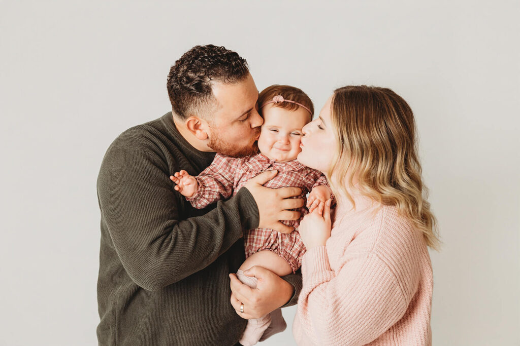 Mom and dad give baby girl a smooch on a simple white backdrop in their studio family photos in mechanicsburg for baby's first birthday