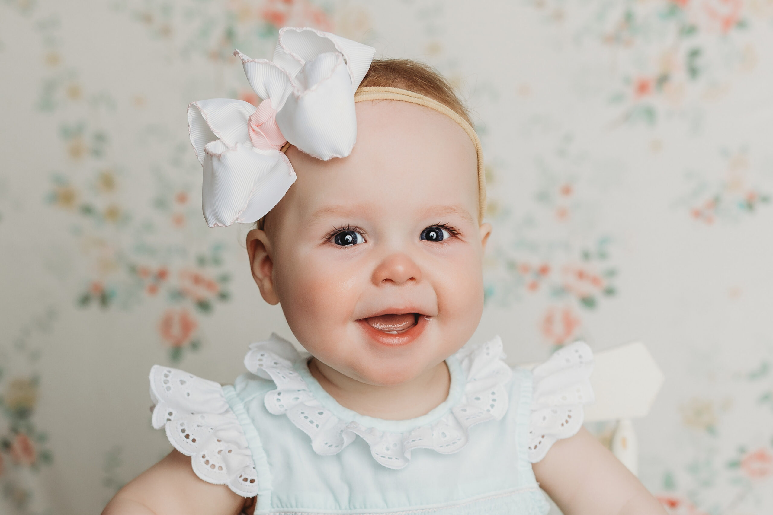 Dillsburg sitter session smily baby girl sitting on a floral backdrop with a big bow and a sweet lacy romper