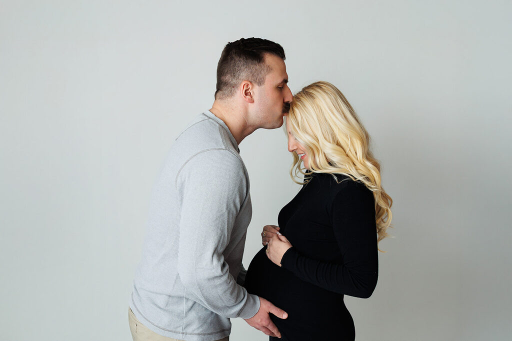 Husband kissing pregnant wife’s forehead during Dover studio maternity photography session