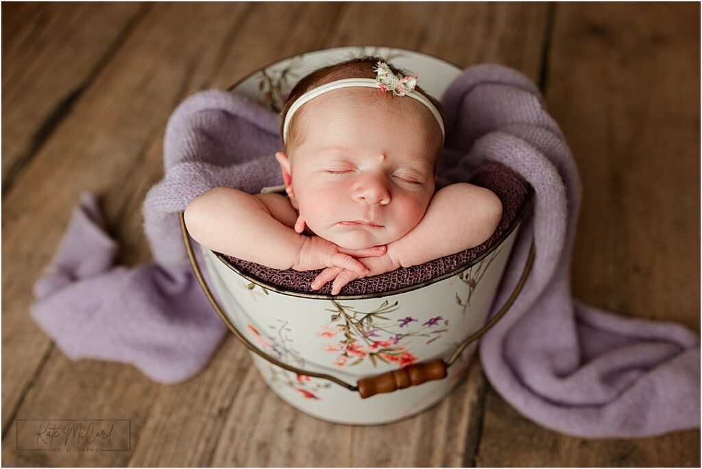 Harrisburg newborn studio photography portrait of baby girl sleeping peacefully in a vintage bucket prop with soft purple wrap, capturing timeless newborn studio portrait.