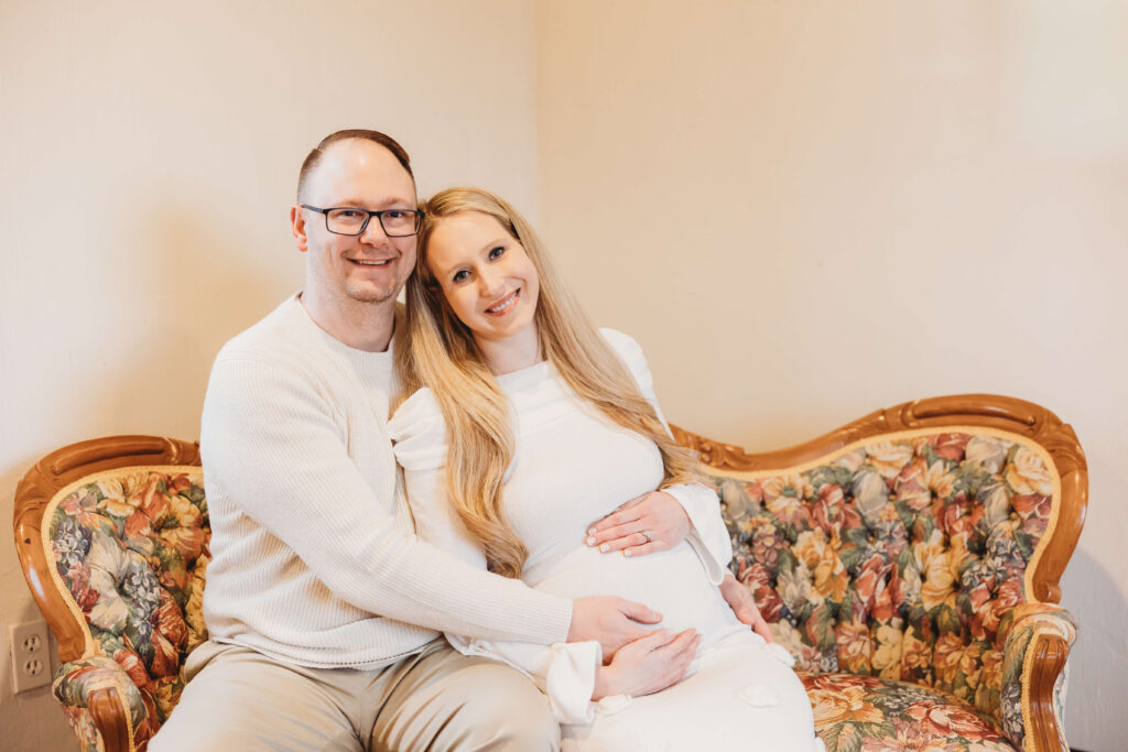 Smiling couple seated on a vintage floral sofa, embracing during their maternity session at Historic Shady Lane, celebrating baby number two.
