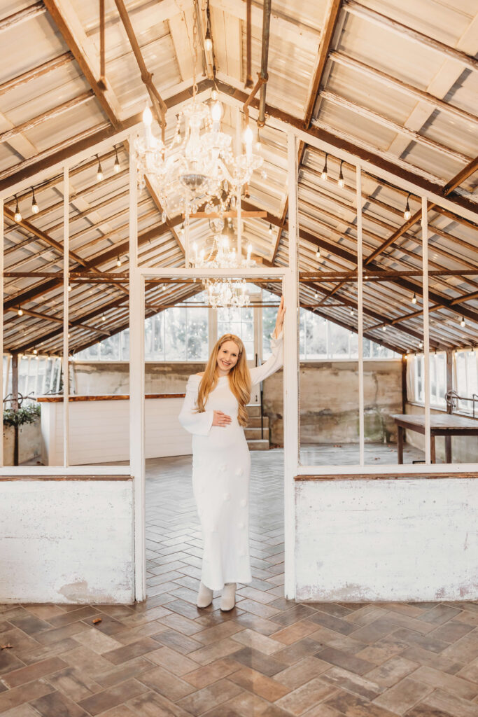 Expectant mother standing inside the greenhouse during her maternity session at Historic Shady Lane, framed by white window panes and glowing chandeliers in Manchester, PA.