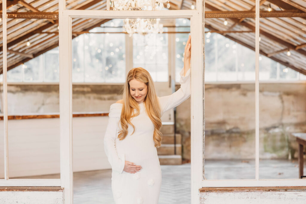 Close portrait of pregnant mother in a soft white dress gently holding her belly inside the Historic Shady Lane greenhouse during a winter York maternity session.