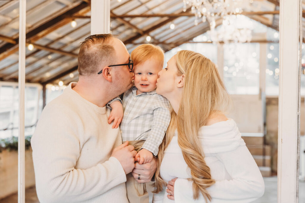 Parents kissing their toddler’s cheeks inside the glowing greenhouse during their maternity session at Historic Shady Lane, capturing a joyful winter York maternity session before welcoming baby number two.