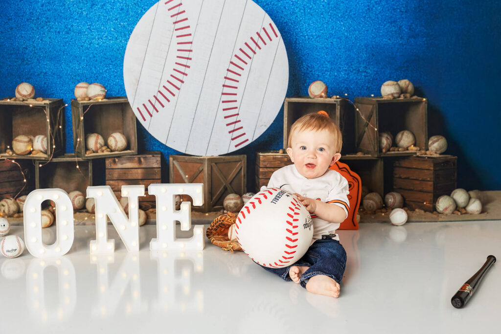 One year old baby boy Orioles baseball cake smash York PA holding large baseball prop with custom themed backdrop