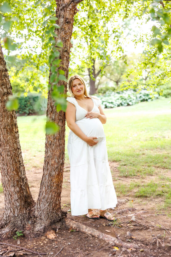 Pregnant mother in a flowing white dress cradling her baby bump beside a tree in a lush garden.