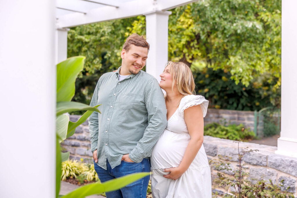Expecting couple smiling together beneath a white pergola during their Hershey Gardens maternity photos.