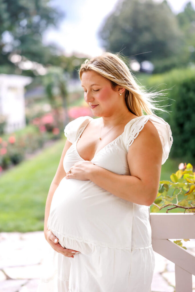 Pregnant mother cradling her baby bump in a white dress during a bright summer maternity portrait.