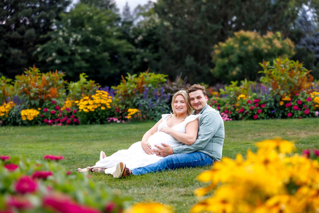 Expecting couple sitting together on the grass surrounded by colorful summer flowers during their Hershey Gardens maternity photos.