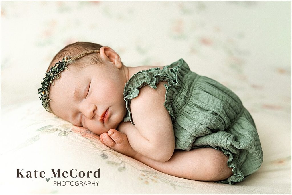 Sleeping newborn baby girl wearing a soft sage green romper and delicate floral headband during a studio newborn session.